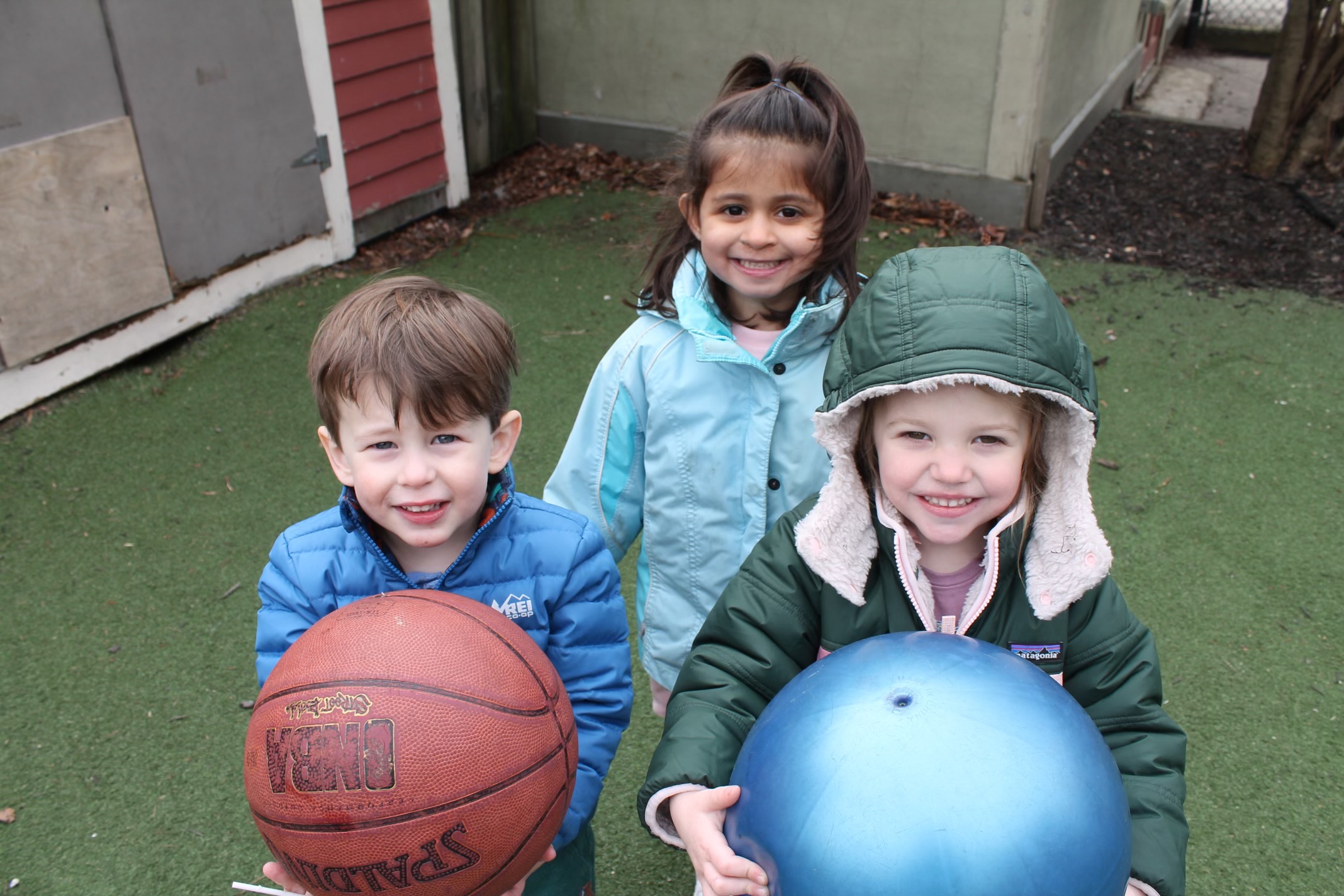 Three children outside at USES in jackets holding a basketball and bouncy ball, looking up at the camera and smiling