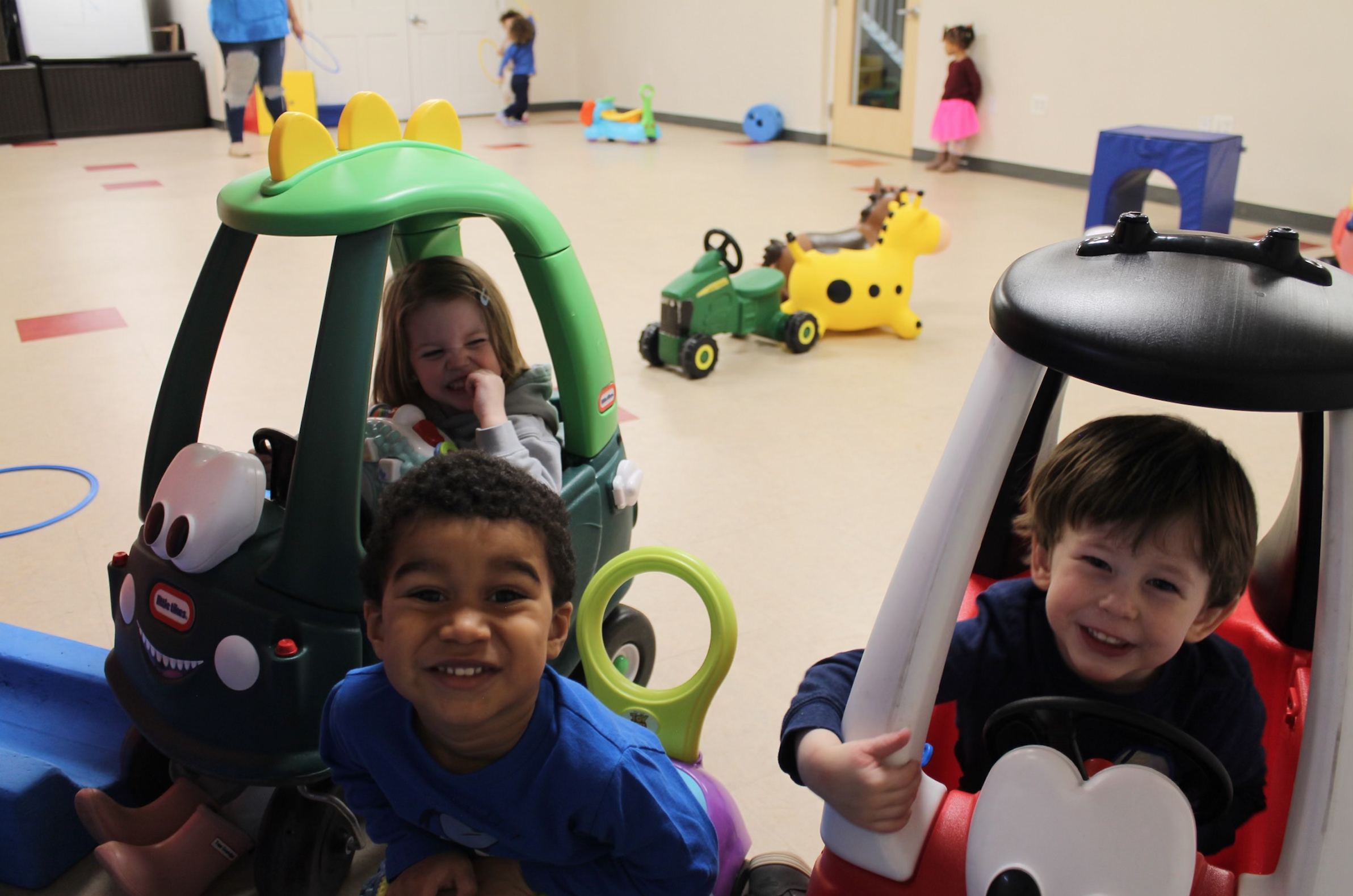 Three children in the USES early childhood education program playing in toy cars and smiling at the camera